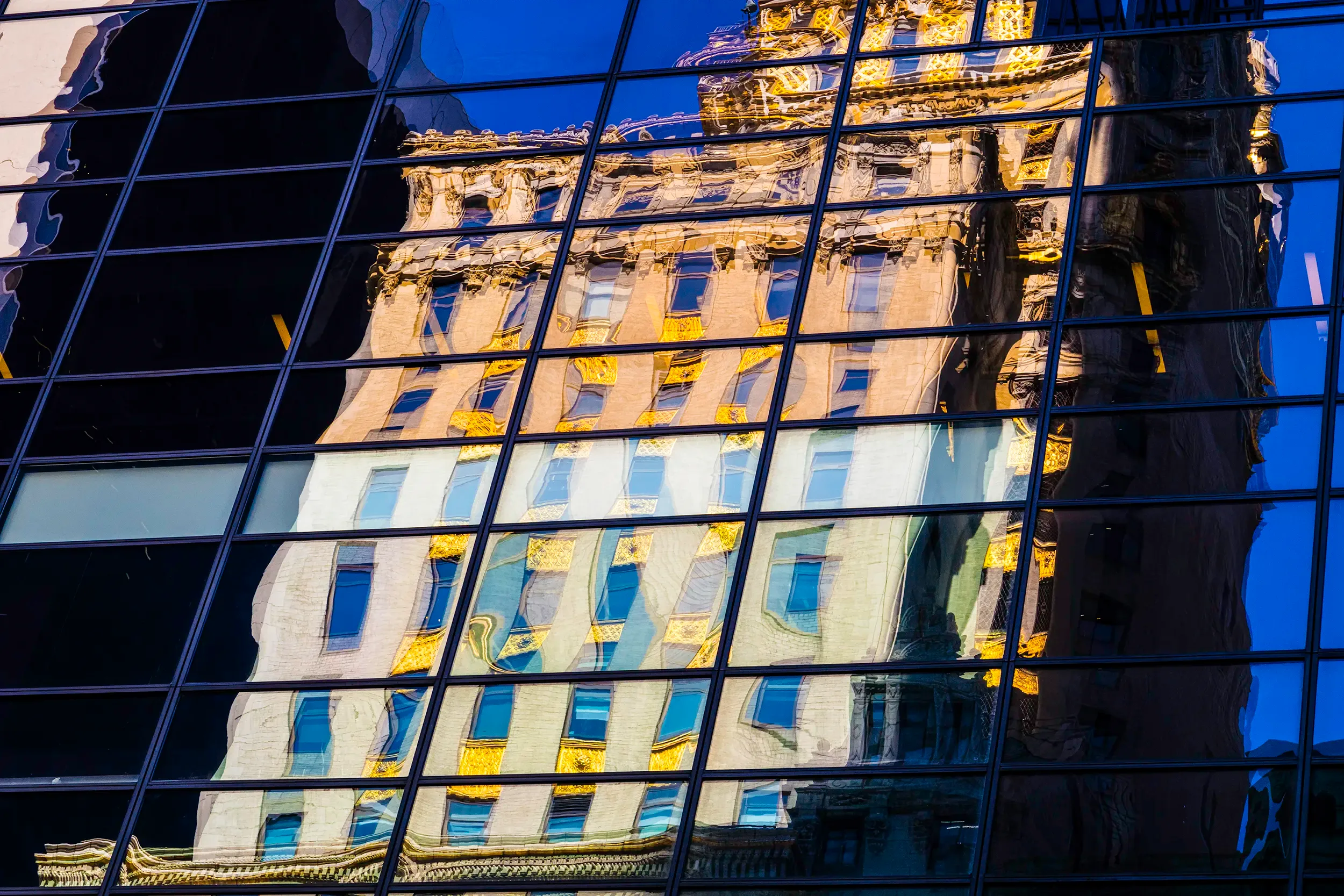 Reflet déformé d’un bâtiment ancien aux ornements dorés sur la façade vitrée d’un immeuble moderne à New York, créant une composition abstraite et colorée entre lignes de verre et détails historiques.