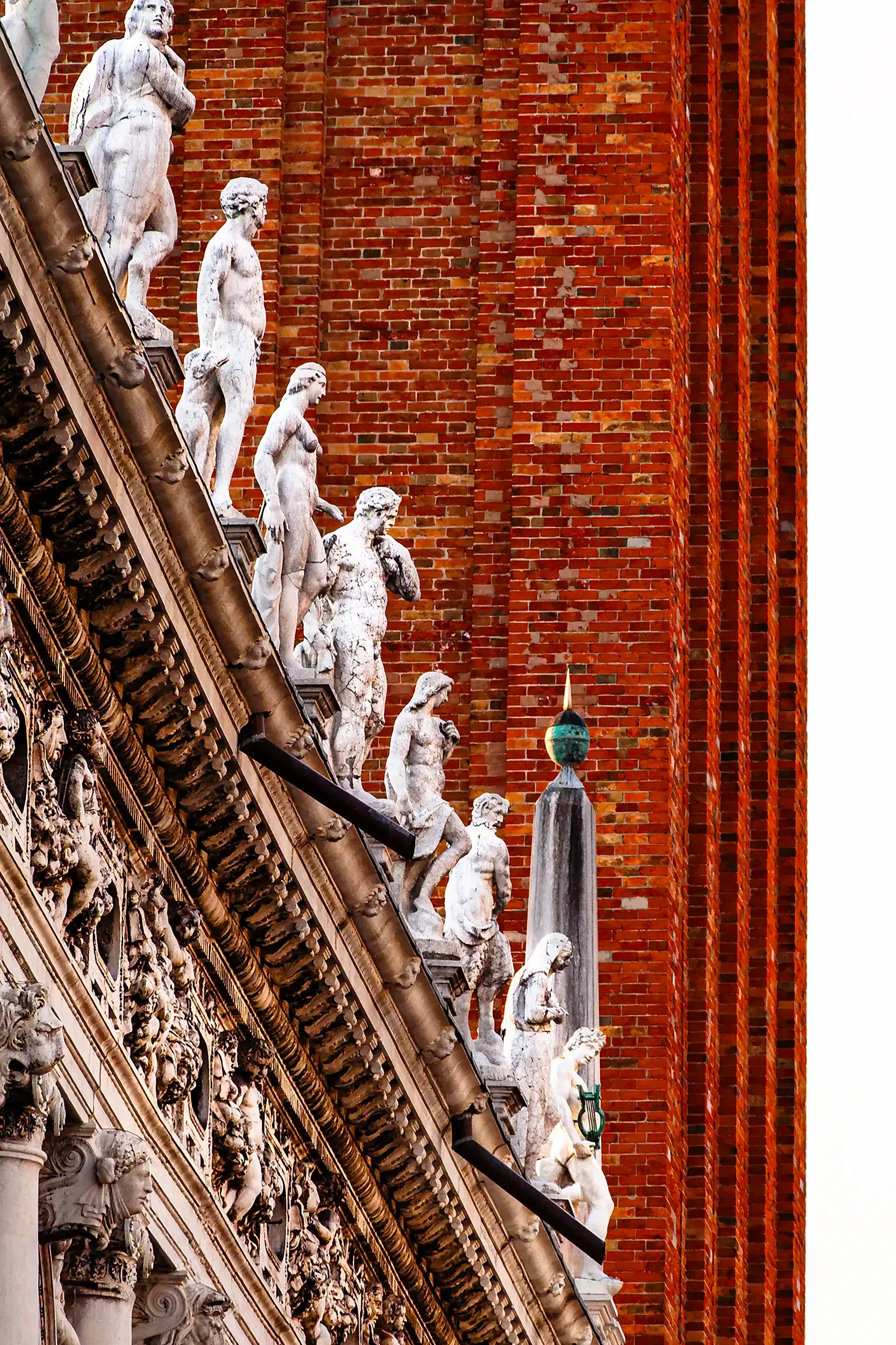 Détail architectural du Palais des Doges à Venise, montrant une frise de statues en marbre blanc se découpant sur le mur de briques rouges du campanile de Saint-Marc. La composition met en valeur la finesse de la sculpture renaissance et le contraste entre la pierre claire et la texture chaude du fond. Une vision élégante et graphique de l’art vénitien.