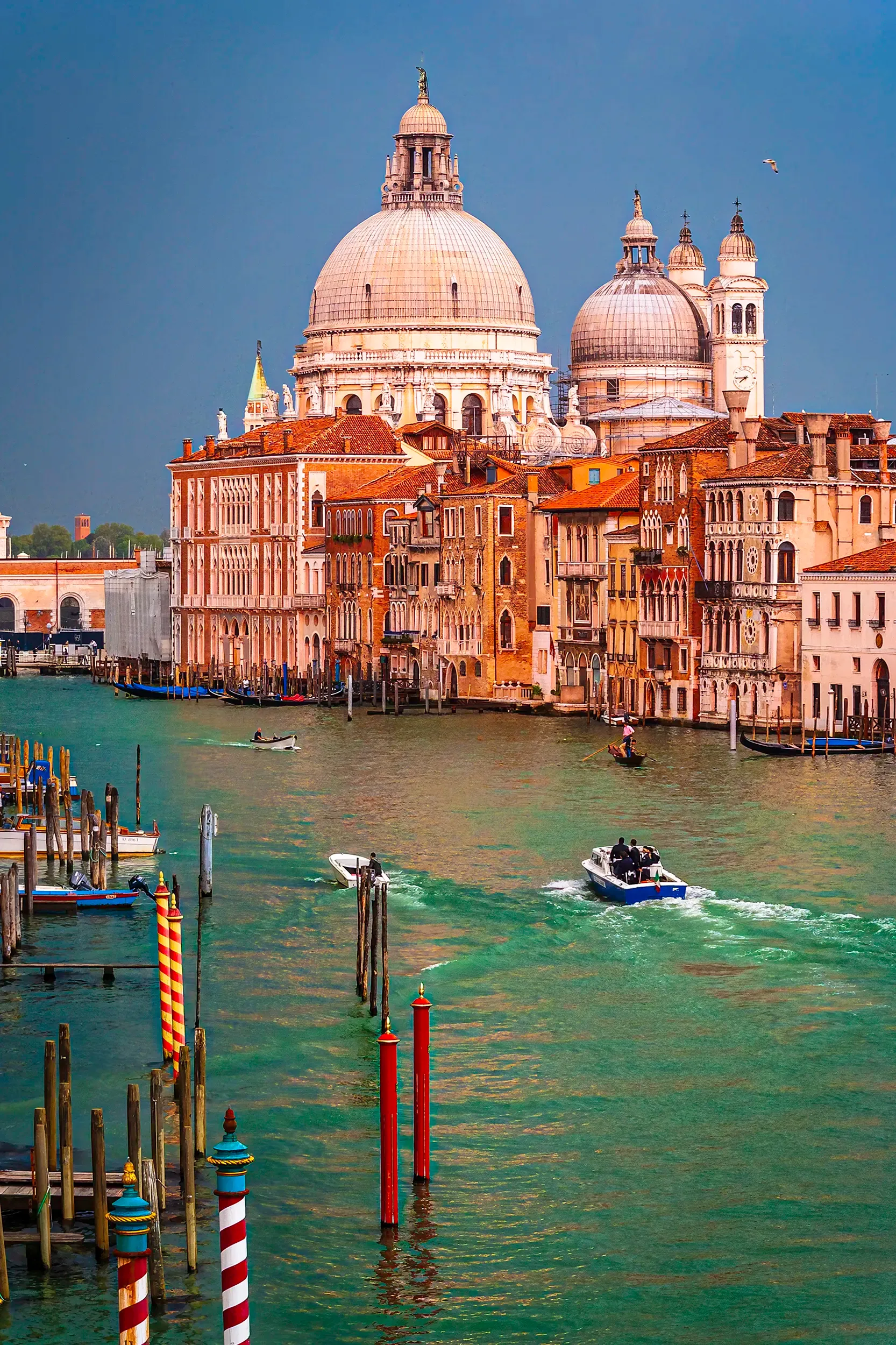 Vue du Grand Canal à Venise dominée par la basilique Santa Maria della Salute, éclairée par une lumière dorée contrastant avec un ciel d’orage bleu profond. Les façades des palais vénitiens se reflètent dans l’eau verte, tandis que des gondoles et bateaux animent la scène. Cette composition met en valeur la grandeur architecturale et l’atmosphère changeante de la lagune vénitienne.