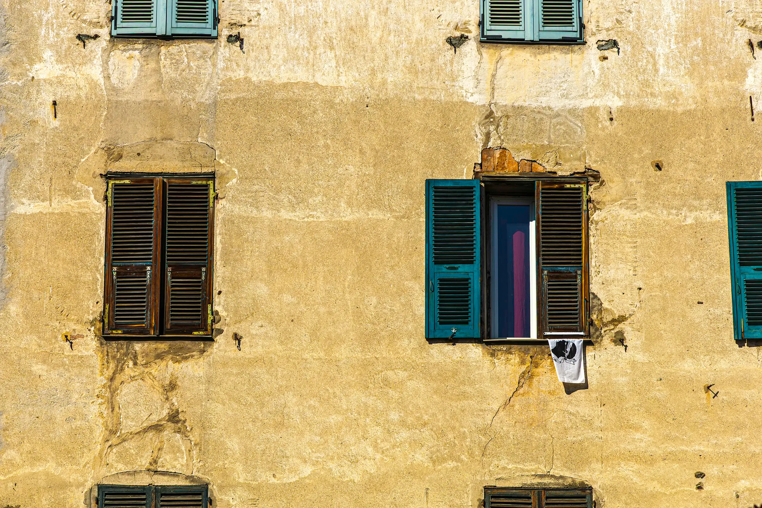 Façade ancienne d’une maison corse avec volets en bois turquoise et bruns, murs beige patinés par le temps, fissures et traces d’usure. Une serviette blanche ornée de la tête de Maure, symbole de la Corse, pend à l’une des fenêtres ouvertes. Photographie mettant en valeur l’architecture traditionnelle corse, la texture des enduits anciens et l’atmosphère méditerranéenne des villages du golfe de Porto et de l’ouest de l’île.